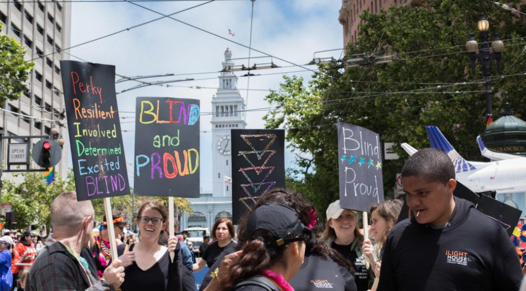 A group of Pride participants gathers near San Francisco’s Ferry Building, holding colorful handmade signs that read “Blind and Proud” and “Blind Proud.”