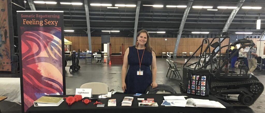 Laura Millar stands behind a LightHouse exhibit table with red tactile anatomy models, books, brochures, and condoms.