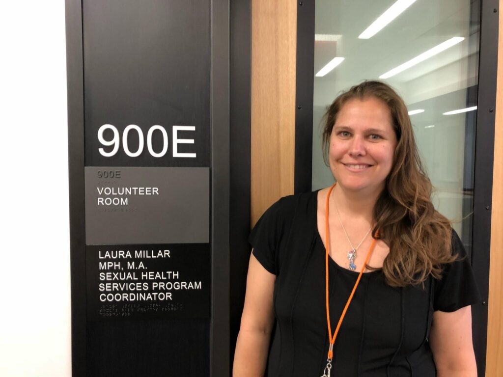 Laura Millar stands beside a doorway sign bearing her name and the title Sexual Health Services Program Coordinator.